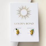 A pair of gold stud earrings with lemon-yellow stones and green leaf accents, presented on a white card with 'GOLDEN BOND' written at the top.